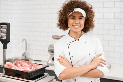 Front view of cheerful girl posing with crossed arms behind glass counter with fresh meat products. Portrait of young smiling woman with curly hair looking at camera, meat in bowl on scales.