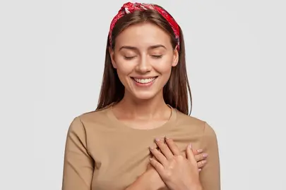 Honest kind hearted woman keeps both palms on chest, expresses kindness, keeps eyes shut, dressed in casual outfit, isolated over white background. People, feeling and body language concept.