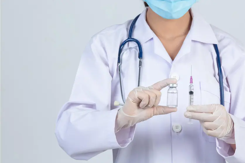 Young doctor is  holding  hypodermic syringe with  vaccine vial and rubber gloves on gray background.