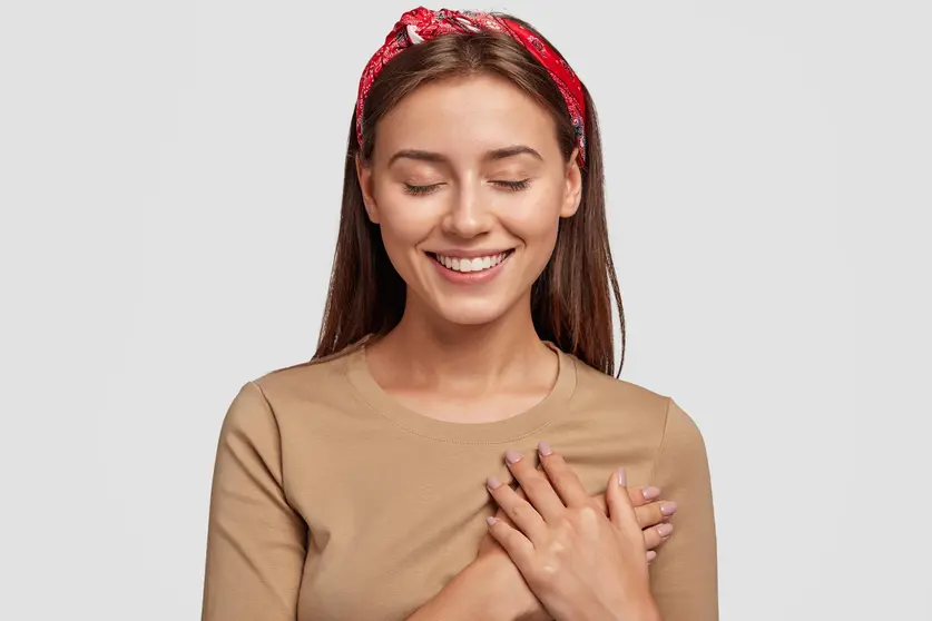 Honest kind hearted woman keeps both palms on chest, expresses kindness, keeps eyes shut, dressed in casual outfit, isolated over white background. People, feeling and body language concept.