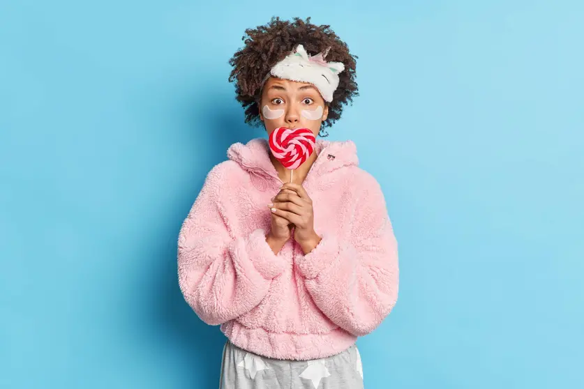 Surprised dark skinned woman has curly hair covers mouth with delicious candy wears sleepmask and pajama applies collagen patches under eyes to reduce wrinkles isolated over blue background.