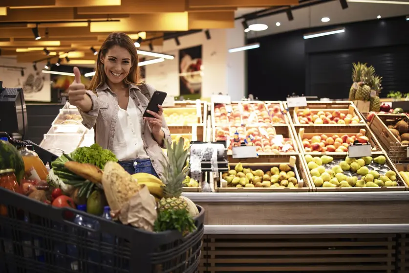 Woman with smart phone in supermarket standing by the shelves full of fruit at grocery store holding thumbs up.