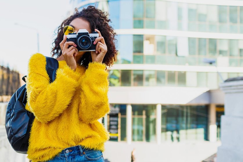  black female photographer making photos on modern architecture background. Wearing back pack, yellow sweater and glasses.