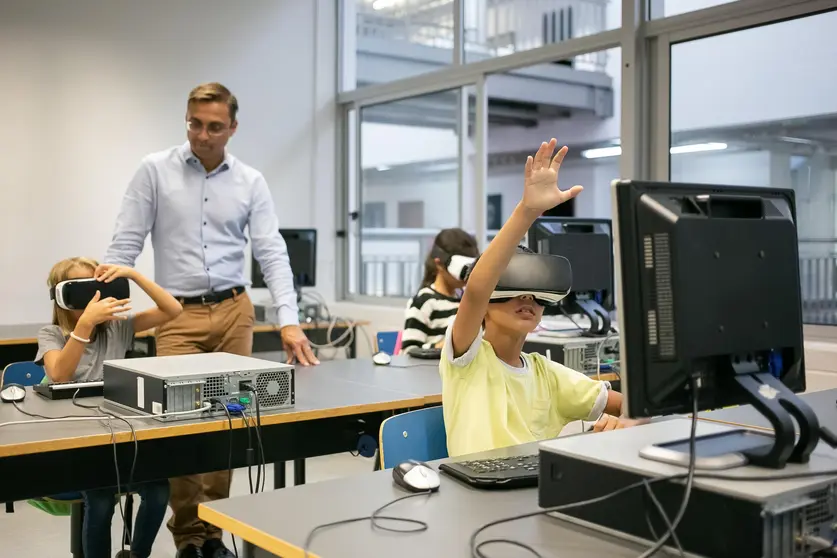 Group of multiethnic kids wearing VR headsets and teacher watching them. Adorable mixed-raced children learning virtual reality and sitting at computer lesson. Informatics and education concept