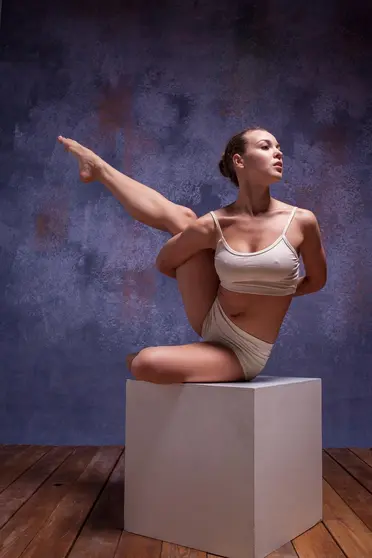 Young beautiful dancer in beige swimwear posing on white cube on lilac studio background 